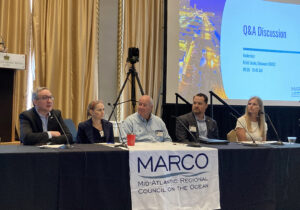 A panel of five seated members speak to the audience